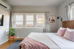 Um quarto com uma cama, uma janela com persianas, uma planta e um quadro na parede.
