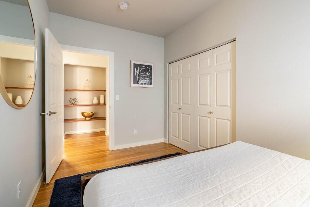 Un dormitorio con cama, armario, espejo, estante con adornos y puerta que da a otra habitación.
