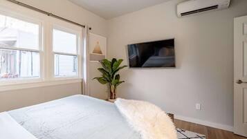 Un dormitorio con cama, televisor, aire acondicionado, ventana y una planta.