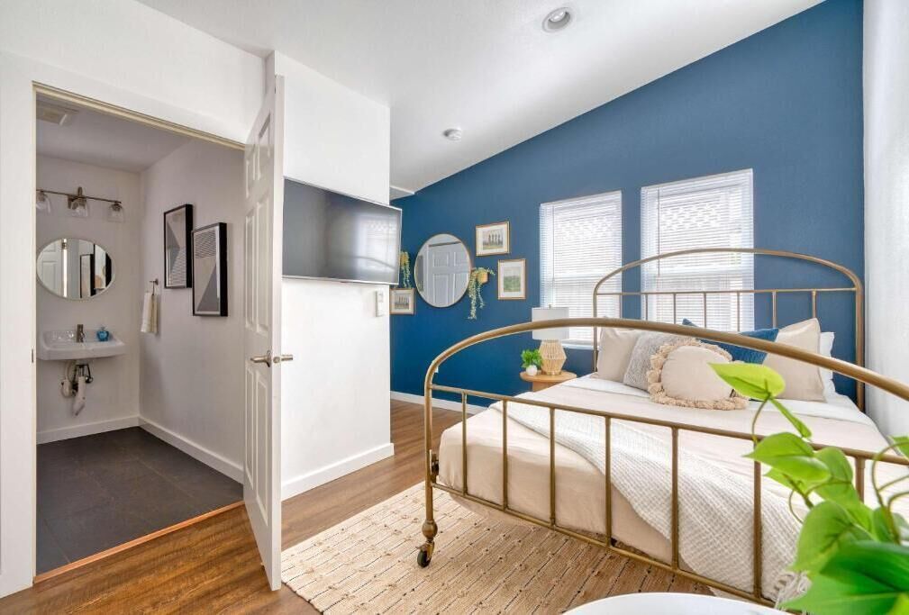A bedroom with a bed, a bedside table, a mirror, and a window with blinds.