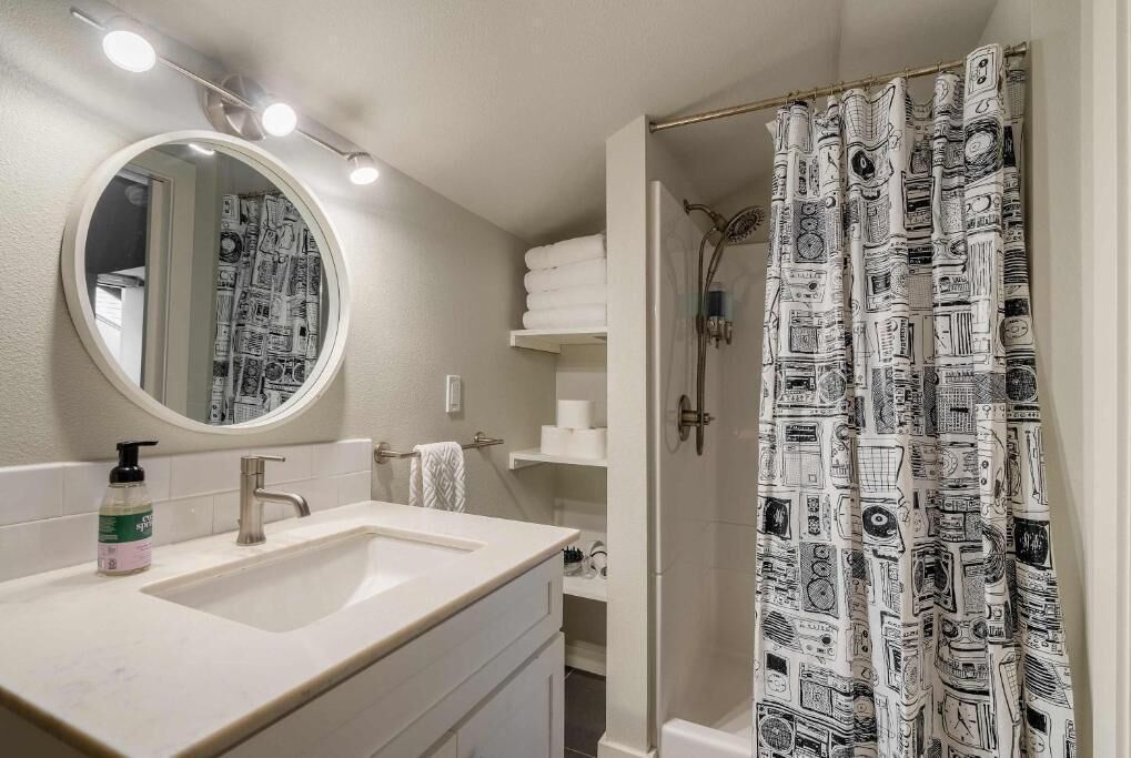 A bathroom with a double sink, a mirror, a shower curtain with a pattern, and a shelf with towels.