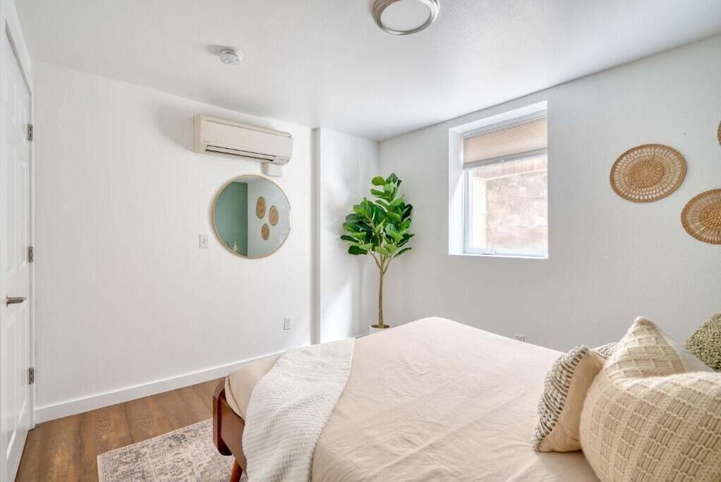 A bedroom with a bed, a round mirror, a potted plant, a window, and a wall-mounted air conditioner.