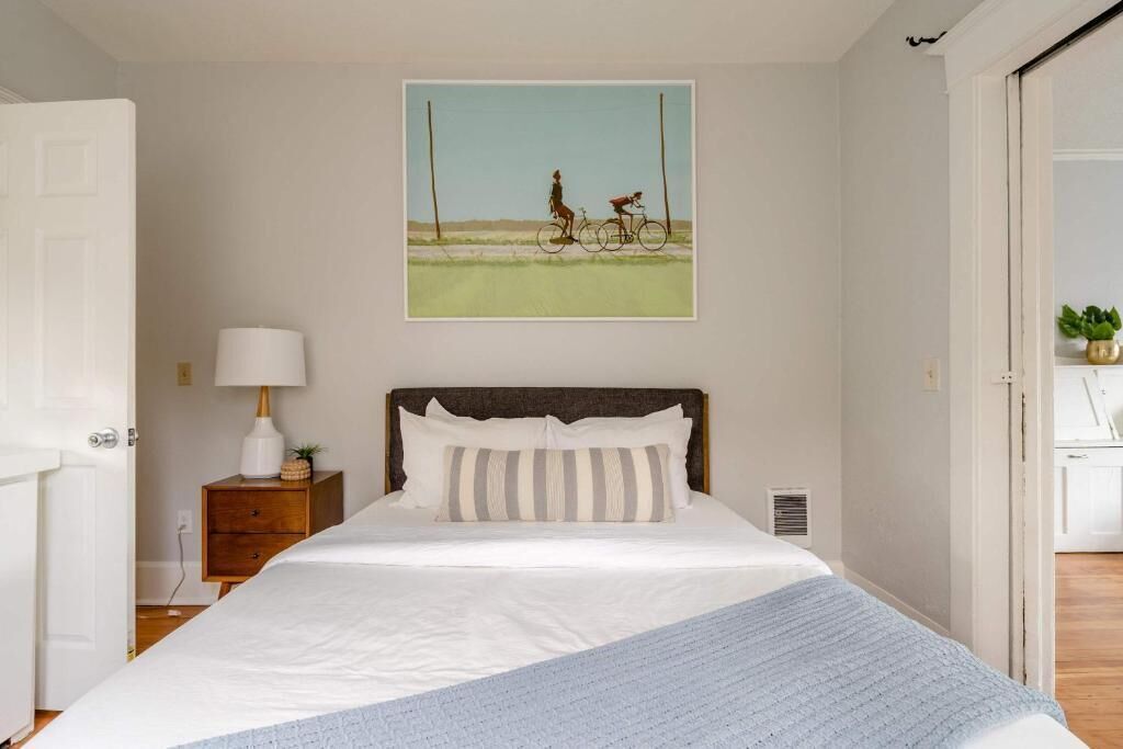 A bedroom with a bed, a nightstand, a lamp, a framed picture on the wall, and a view of the outside.