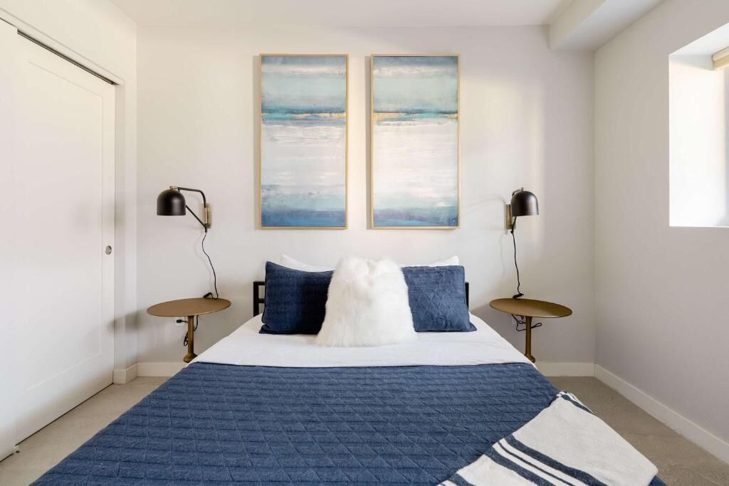 A bedroom with a bed, two wall-mounted paintings, two bedside lamps, and a window.