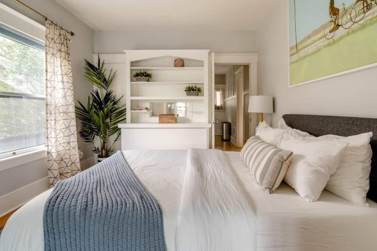 Un dormitorio con cama, mesita de noche, lámpara, ventana con cortinas, una planta y estante con objetos.