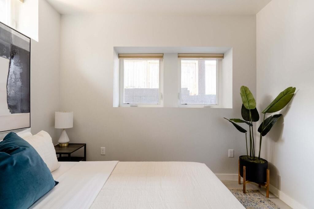 Un dormitorio con una cama, una mesita de noche, una lámpara, dos ventanas y una planta en maceta.