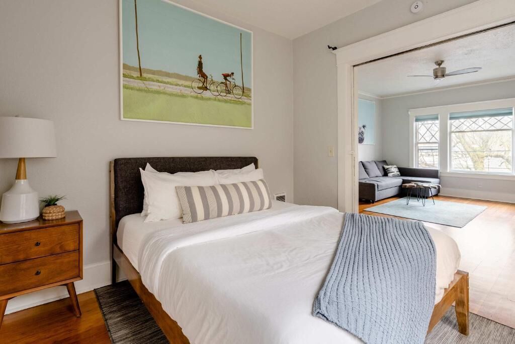 A bedroom with a bed, a nightstand, a lamp, a painting, a window, and a ceiling fan.