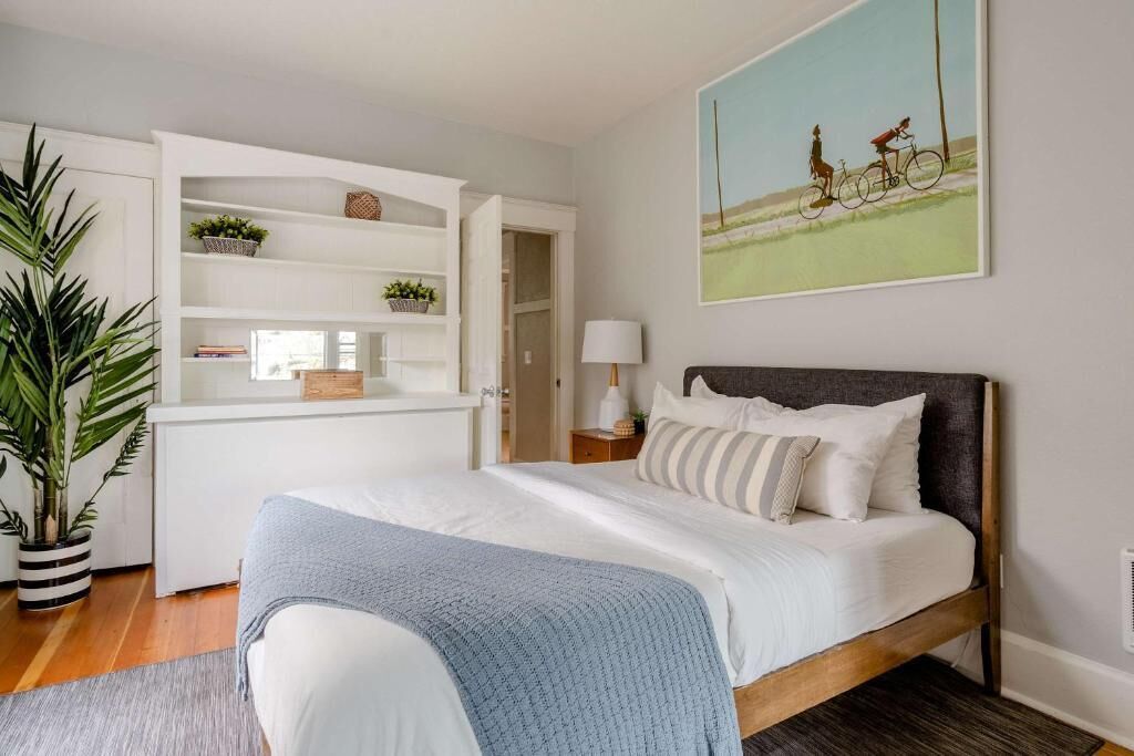A bedroom with a bed, a white wardrobe, a potted plant, a painting, and a nightstand with a lamp.