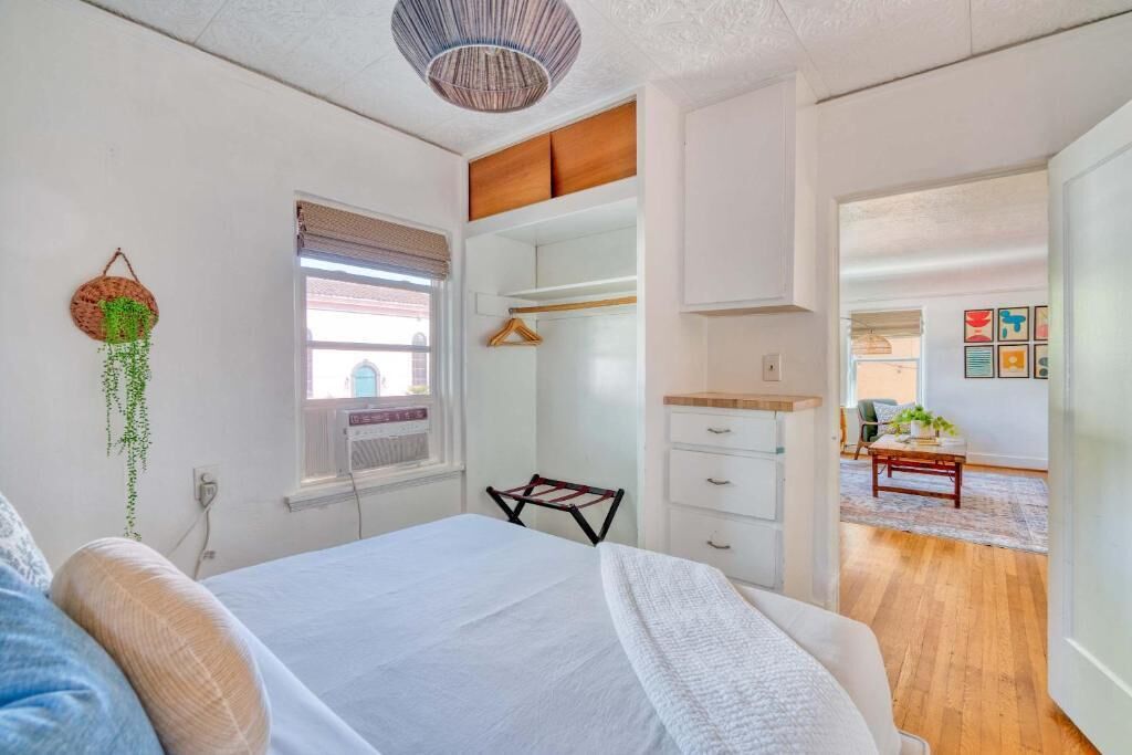 A bedroom with a bed, a hanging plant, a window with blinds, a wardrobe, and a view of another room.