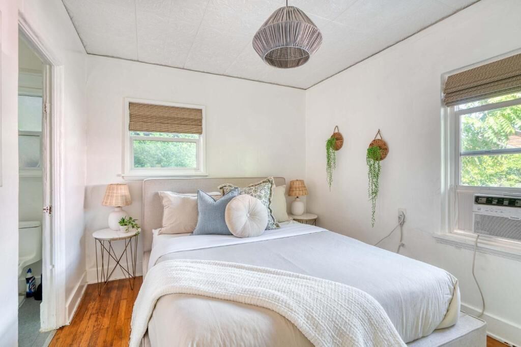 A bedroom with a bed, bedside tables, a hanging plant, a window with blinds, and a ceiling light fixture.