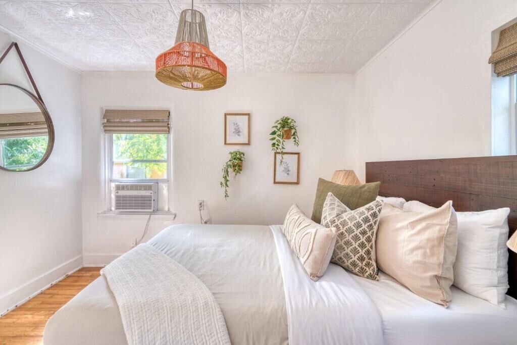 A bedroom with a large bed, a hanging light fixture, a window with blinds, and wall-mounted plants.