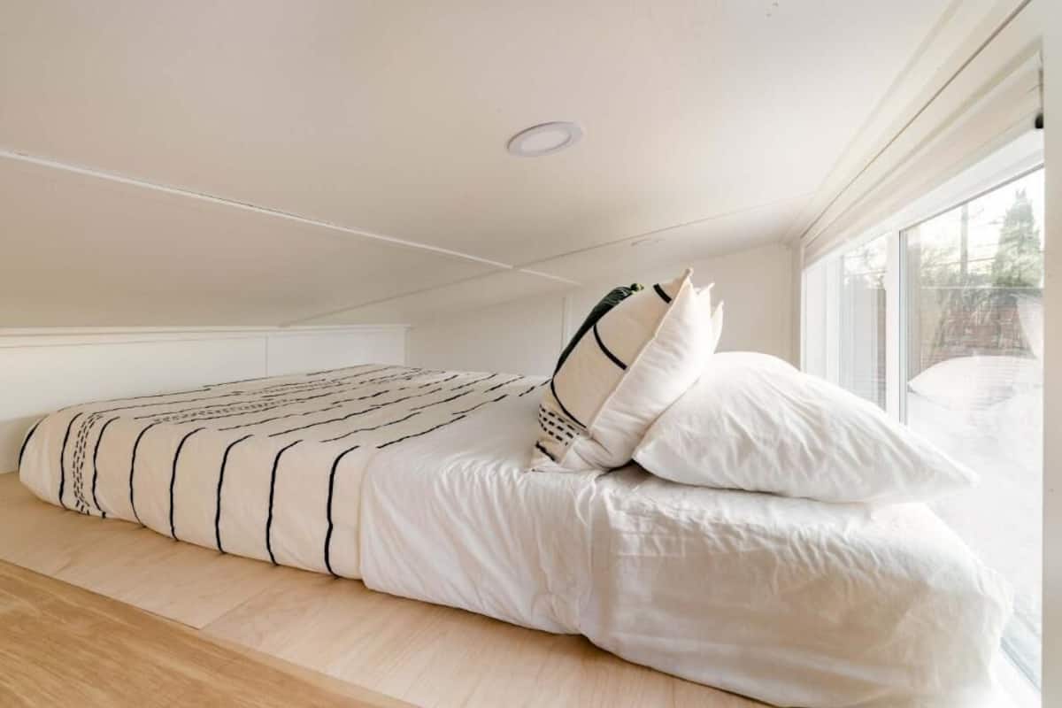 Una cama individual con una colcha estampada y almohadas, suelo de madera y ventana con vistas a árboles.