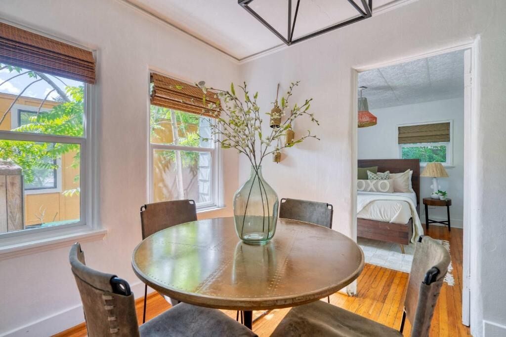 A dining area with a round table, chairs, a vase with branches, and a bed in the background.