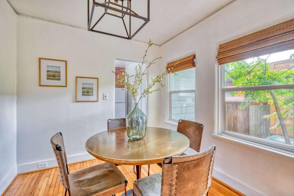 Un comedor con una mesa redonda, sillas, un florero con ramas, cuadros en la pared y una ventana con vista a la vegetación.