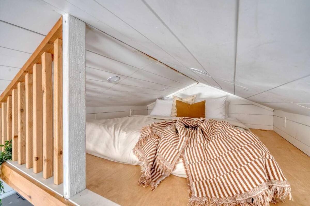 Un dormitorio acogedor en el ático, con techo inclinado, barandilla de madera, cama con colcha a rayas y ventana que proporciona luz natural.