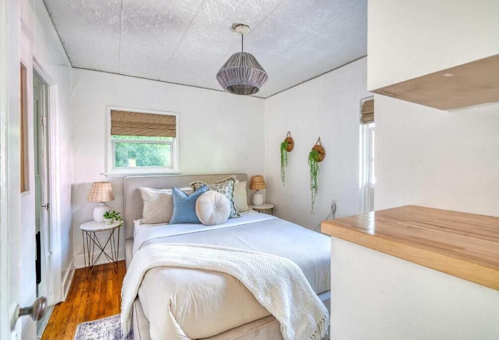 A bedroom with a bed, bedside tables, a window with blinds, a hanging plant, and a ceiling light fixture.