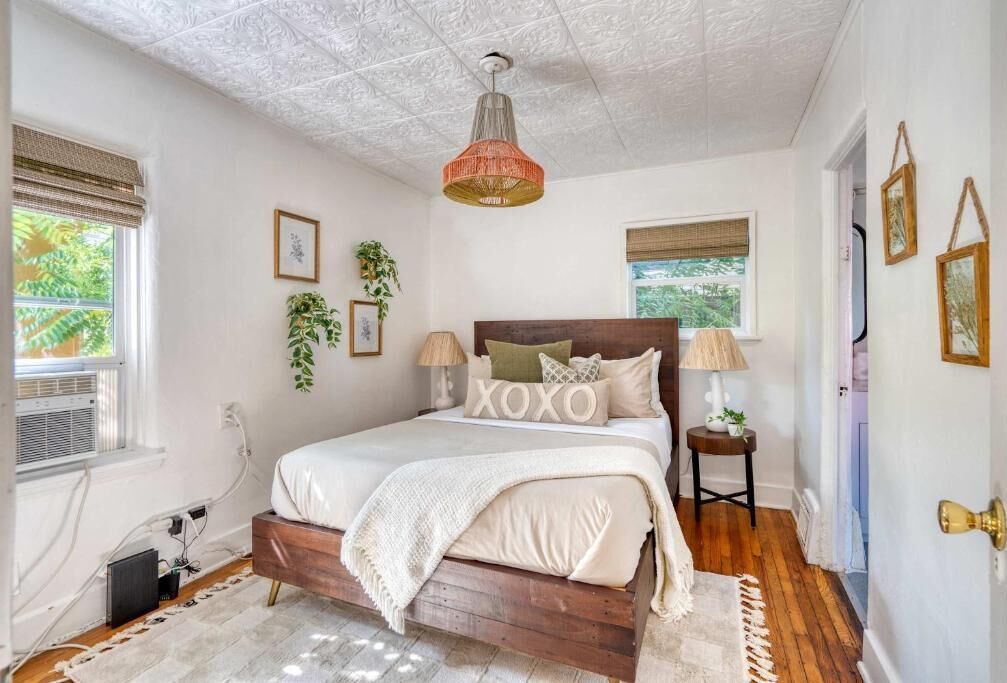 Un dormitorio con cama de madera, ventilador de techo, ventana con persianas y una planta colgada en la pared.