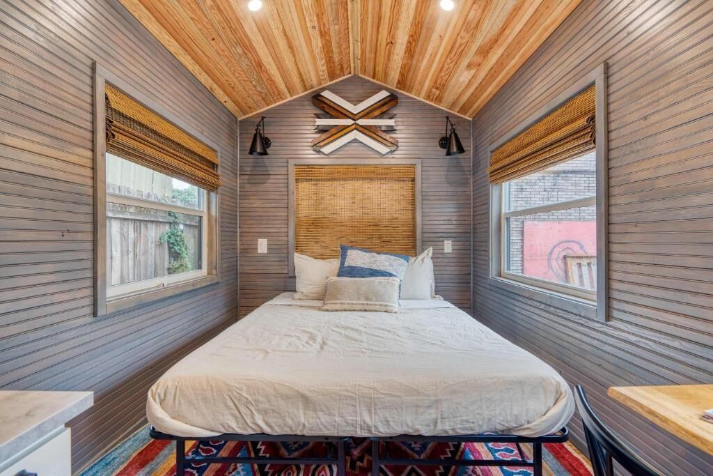 Una habitación estrecha con una cama, techo de madera y dos ventanas con persianas.