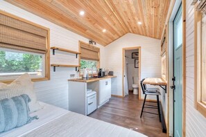 A compact, well-lit room with a wooden ceiling, a kitchenette, a bed with pillows, and a view through a window.