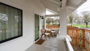 A covered porch with a wooden deck, a green door, and a view of a park.