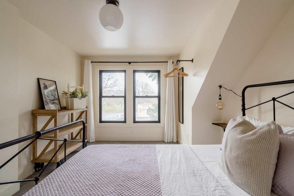 A bedroom with a bed, a bedside table, a hanging light fixture, and a window with curtains.