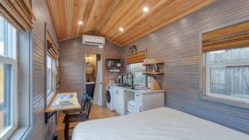 A compact room with a bed, a small kitchenette, a washing machine, and a dining area with a table and chairs.