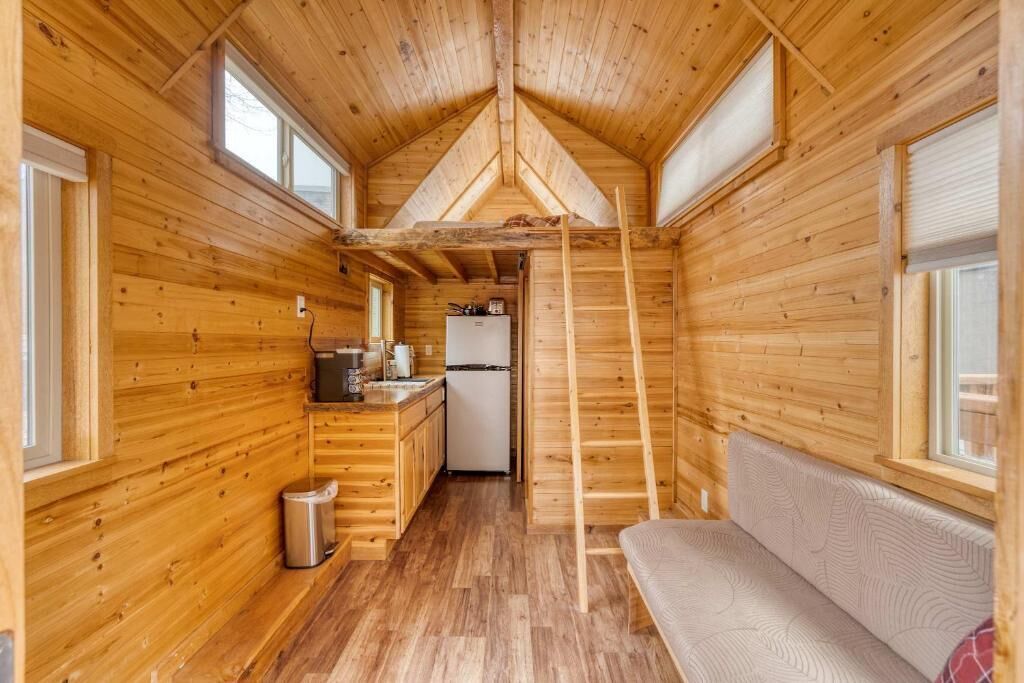 A compact wooden cabin with a kitchenette, a sofa, and a ladder.