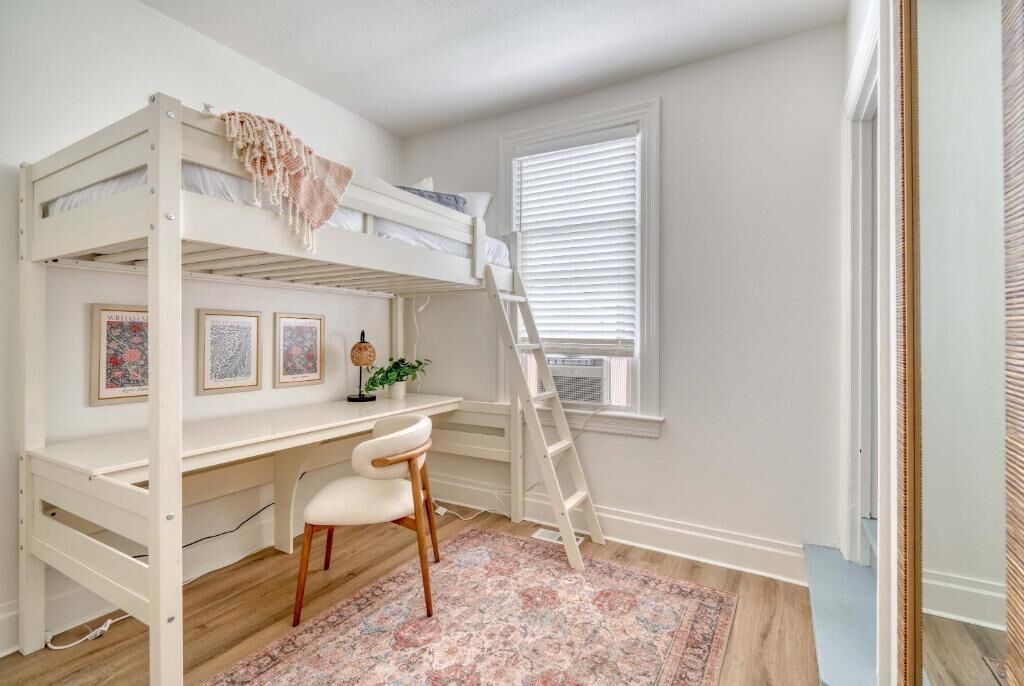 A bunk bed with a desk and chair, a ladder, framed pictures, a plant, and a window with blinds.