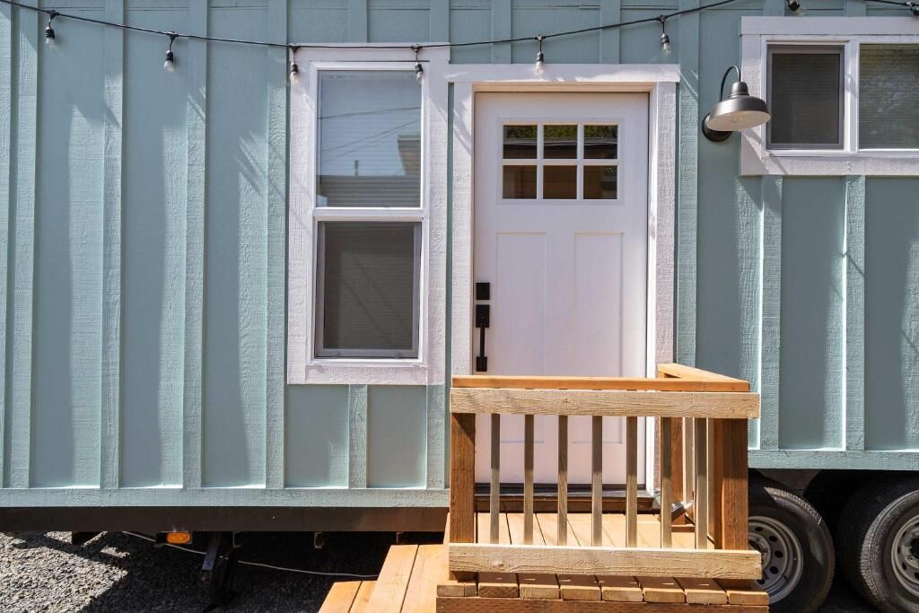 A small, single-story building with a white door and two windows, a wooden deck in front, and a string of lights above.