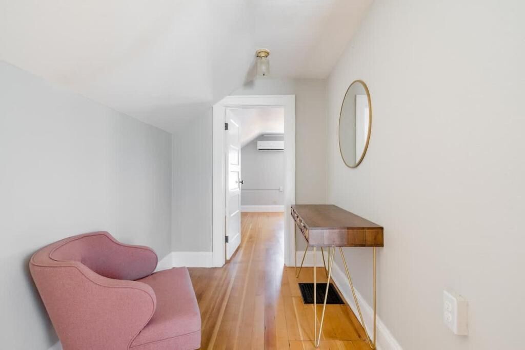 A hallway with a wooden floor, a pink chair, a small table, and a mirror on the wall.