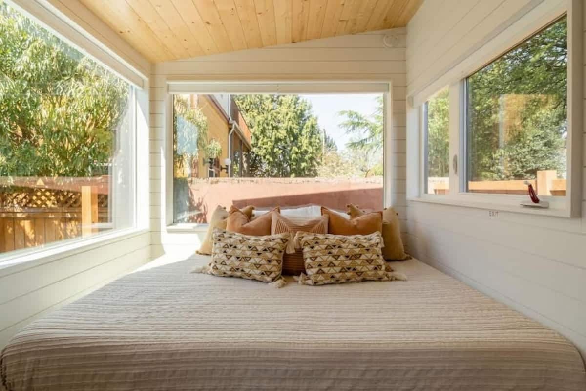 A bedroom with a large bed, multiple pillows, a wooden ceiling, and large windows.