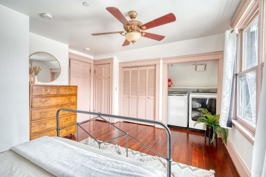 Un dormitorio con una cama, un aparador de madera, un ventilador de techo, una ventana y una kitchenette.