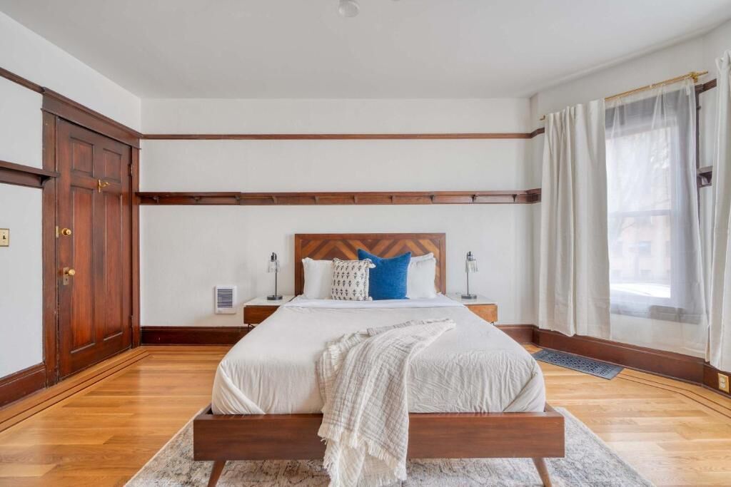 A bedroom with a wooden bed frame, white bedding, a headboard, bedside tables, and a window with curtains.