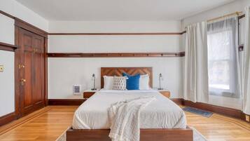 A bedroom with a wooden bed frame, white bedding, a headboard, bedside tables, and a window with curtains.