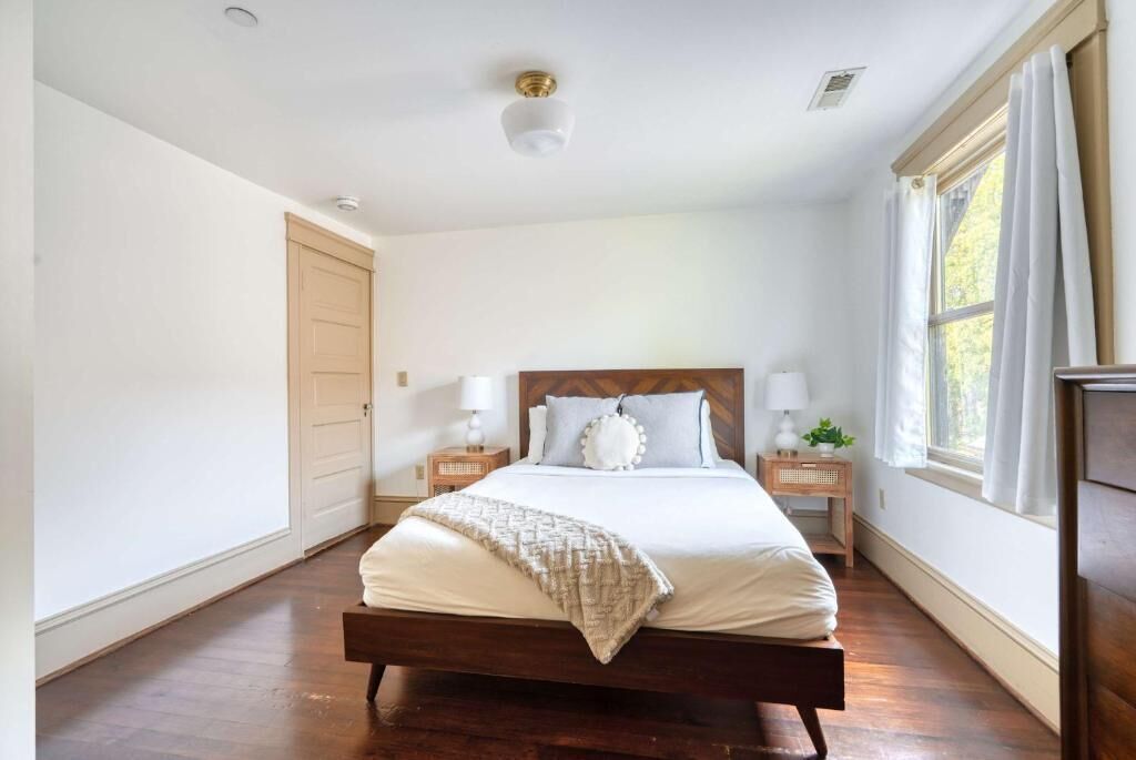 A bedroom with a wooden bed, white walls, a wooden floor, a ceiling light, a window with curtains, and a nightstand with a lamp.