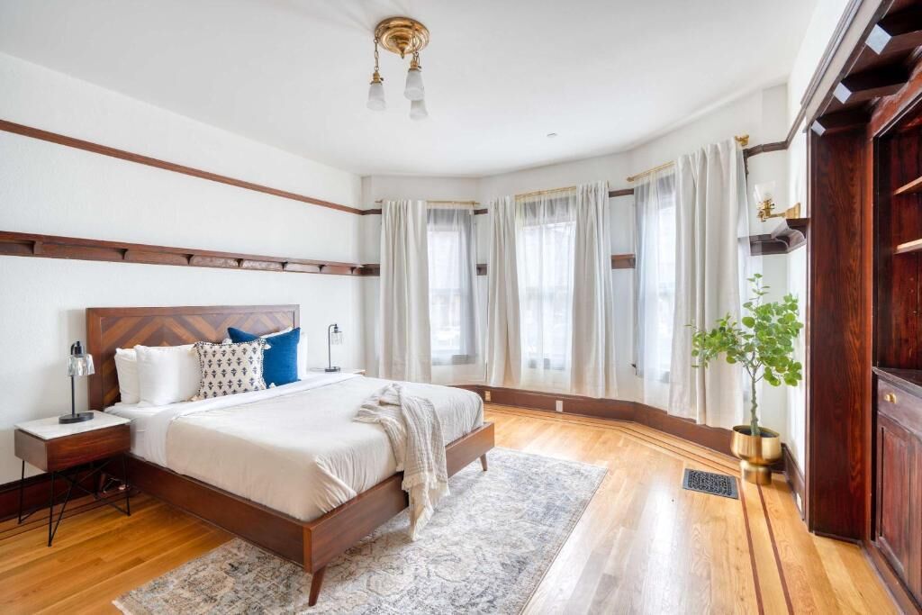 Une chambre avec un lit en bois, du linge de lit blanc, une table de chevet, une lampe de chevet, une grande fenêtre avec des rideaux et une plante en pot.
