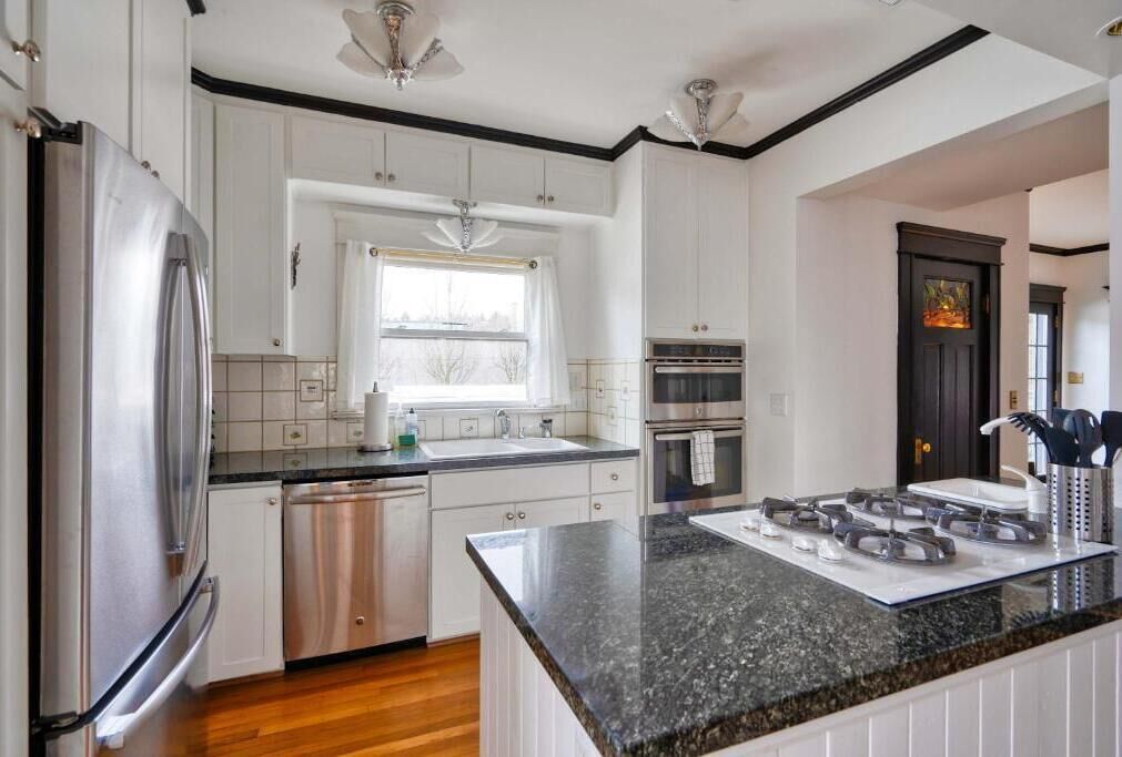 A kitchen with white cabinets, a stainless steel refrigerator, a dishwasher, a window with curtains, a stove, and a black countertop.