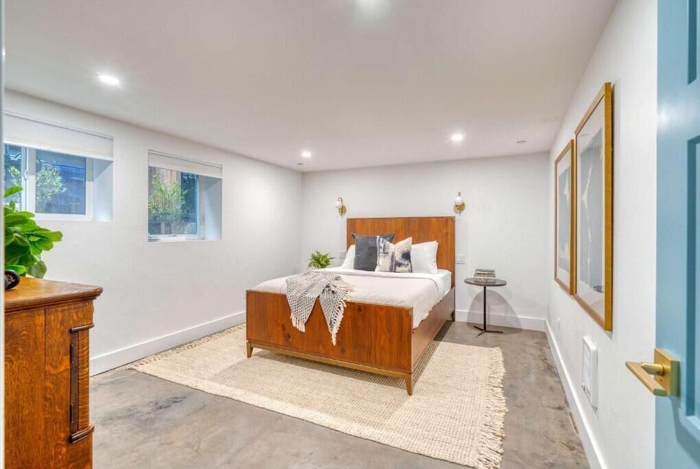 A bedroom with a wooden bed, a small bedside table, a wall-mounted light, and a window.