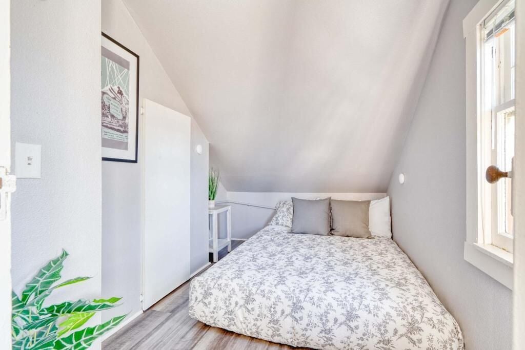 A small, narrow room with a sloped ceiling, a bed with a floral bedspread, a small table with a plant, and a framed picture on the wall.