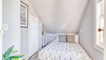 A small, narrow room with a sloped ceiling, a bed with a floral bedspread, a small table with a plant, and a framed picture on the wall.