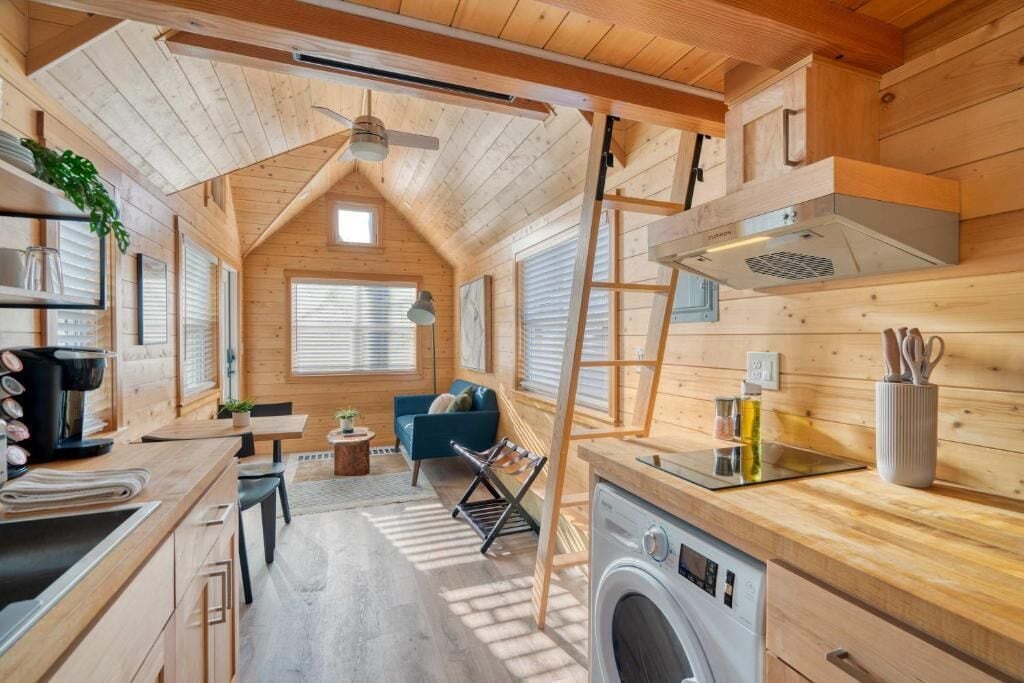 En kompakt, träinredd interiör med ett litet kök, en tvättmaskin, en matplats och en sittgrupp.