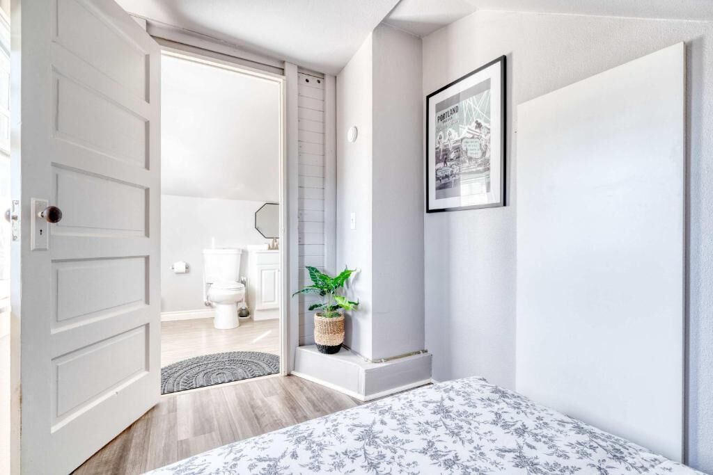 A bedroom with a bed, a door leading to a bathroom, a framed picture on the wall, and a potted plant.