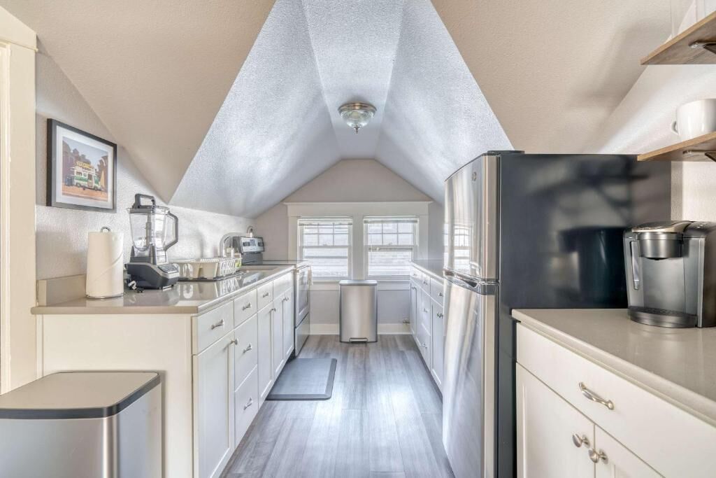 A narrow, modern kitchen with stainless steel appliances, a small trash bin, and a window with blinds.