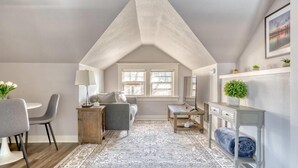Un dormitorio estilo loft con techo inclinado, una zona de comedor con mesa y sillas, una alfombra grande con diseño, y una mesita de noche con lámpara.