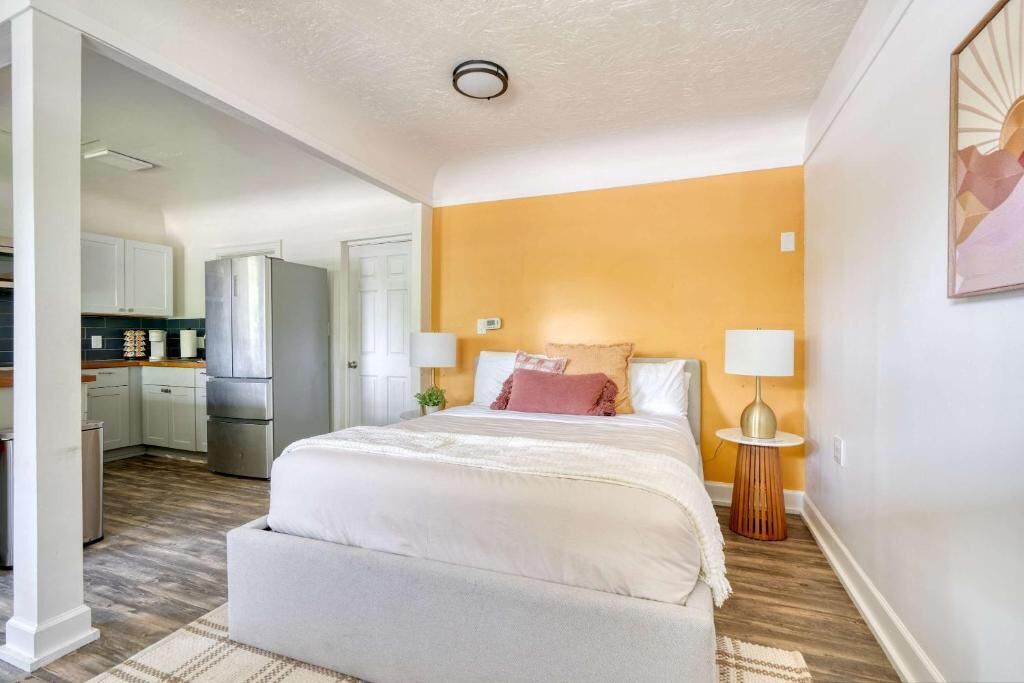 A bedroom with a bed, bedside lamps, a nightstand, and a view into the kitchen.