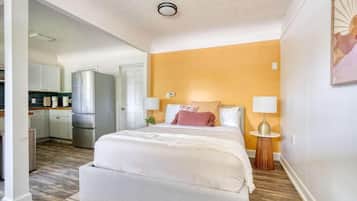 A bedroom with a bed, bedside lamps, a nightstand, and a view into the kitchen.