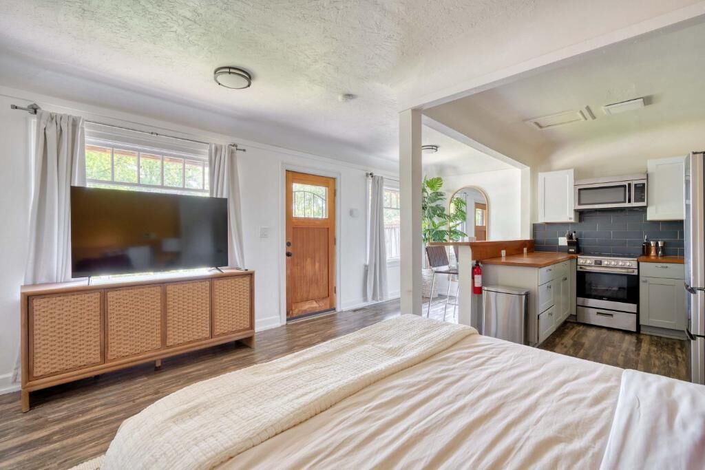 A bedroom with a large bed, a TV, a wooden cabinet, and a kitchen area with a microwave and stove.