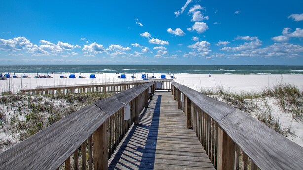 Ein hölzerner Steg führt zu einem Strand mit blauen Stühlen und blauem, wolkenlosem Himmel.