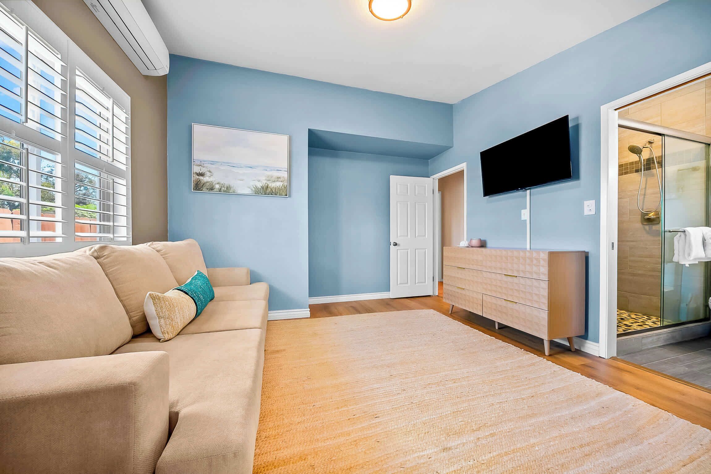 A living room with a beige sofa, a flat-screen TV, a painting, and a glass shower area.
