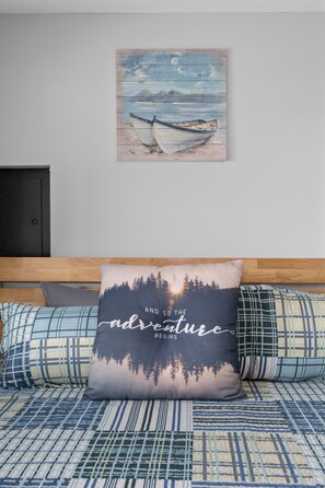 A bedroom with a bed, a pillow with a forest and sunset design, and a painting of boats on a wooden wall.
